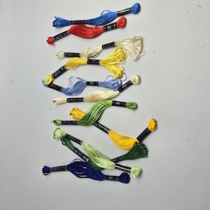 May include: A collection of embroidery floss skeins in various colors including blue, red, cream, yellow, light blue, light green, dark green, and dark blue. Each skein is secured with a black paper band featuring white printed text and numbers.