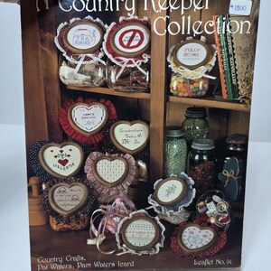 May include: A Country Keeper Collection leaflet with cross-stitch patterns for various home decor items, including tea bags, jelly beans, and welcome signs. The leaflet is from Country Crafts by Pat Waters and Pam Waters leard.