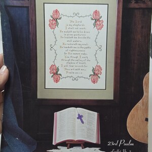 May include: A cross stitch pattern featuring the 23rd Psalm, "The Lord is my shepherd, I shall not want." The pattern is surrounded by pink roses and a green background. The text is in black and gold. The pattern is framed in a brown frame.