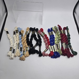 May include: A collection of embroidery floss skeins arranged in a row, featuring a variety of colors including white, light blue, tan, black, pink, red, blue, green, and dark maroon, each secured with a small gold and black paper label.