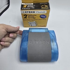 May include: A blue and gray Xyron Cheetah adhesive runner with a matching cartridge box. The box is yellow and black with the text "Xyron Cheetah" and "Adhesive Runner Cartridge". The runner is acid-free and permanent.