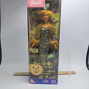 May include: A Barbie doll in a box labeled Halloween Enchantress and La Noche de Brujas. The doll wears a black dress with gold stars, a matching witch hat, and a gold sheer cape. The packaging features a pumpkin graphic and purple bats.