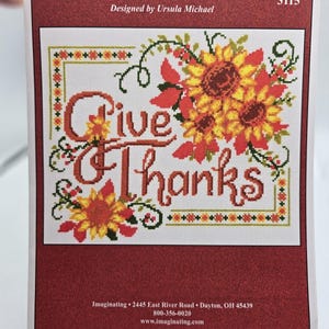 May include: A cross-stitch pattern featuring the words "Give Thanks" in orange script, surrounded by sunflowers and floral accents. The design is set against a white background with a red border. The text "GIVE THANKS SUNFLOWERS" is at the top.