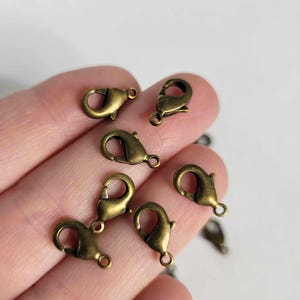 May include: A close-up of a hand holding a group of antique gold-toned metal lobster claw clasps. The clasps are used for jewelry making and crafting.