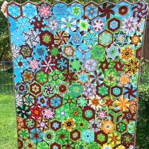 May include: A quilt featuring a vibrant mosaic of hexagonal fabric patches in various colors including blue, green, orange, and brown. The design incorporates diverse patterns such as floral, geometric, and animal prints, finished with a brown scalloped edge.