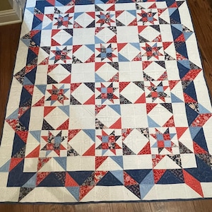 May include: A rectangular quilt featuring a repeating star pattern in shades of red, white, and blue. The design incorporates various floral and geometric fabric prints within the star points, set against a white background with a blue border.