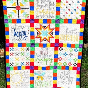 May include: A multi-colored patchwork quilt featuring nine square blocks separated by a rainbow-colored border. The blocks contain various patterns, including a star, a butterfly, and text such as "find your happy place" and "be the sunshine."