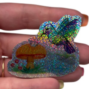 May include: A holographic sticker featuring a hummingbird in flight above a yellow mushroom. The sticker has a reflective, multi-colored glitter finish that catches the light, displaying a vibrant spectrum of colors across the bird and mushroom.