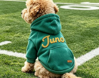 May include: A green dog hoodie featuring a hood, a kangaroo pocket, and the name Juno embroidered in yellow cursive script on the back. The garment includes a small rectangular brown patch on the pocket and ribbed cuffs at the sleeves.