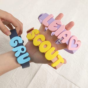 Pode incluir: Três coleiras de silicone com letras coloridas em relevo. Uma coleira azul-escura exibe o nome GRU em azul-claro. Uma coleira cor de pêssego apresenta o nome SCOUT em amarelo. Uma coleira lavanda mostra o nome LEIA com um símbolo de coração.