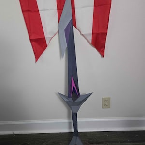 May include: A stylized sword prop featuring a dark gray blade with a light gray pointed tip, a dark gray hilt with a purple triangular accent, and a dark gray handle. The sword has a geometric, angular design with a pointed pommel at the base.