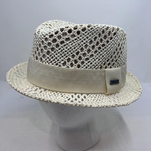 May include: A cream-coloured fedora hat featuring an open-weave straw construction with a wide, solid-coloured fabric band encircling the base. A small, rectangular metallic plaque is attached to the side of the band, displaying the brand name Stetson.