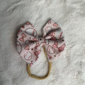 May include: A light pink baby bow headband with a baseball theme. The bow features a pattern of baseballs, bows, and stars. The headband is made of a soft, stretchy material.