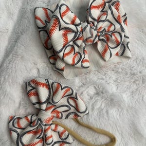 May include: Two baseball-themed baby headbands. One is a wide headband with a large bow, and the other is a smaller bow on a thin, tan elastic band. Both have a white background with a repeating pattern of baseballs in red and black.