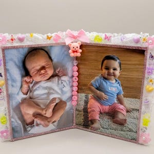 May include: A double-sided hinged picture frame decorated with white lace, pink rhinestones, and various colorful charms including hearts, flowers, and pacifiers. A pink bow and a small bear figure sit at the top, with the name ISABELLA spelled in beads.