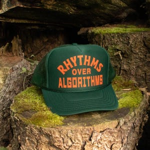 May include: A dark green trucker hat featuring a mesh back and a solid front panel. The text RHYTHMS OVER ALGORITHMS is printed in bold orange capital letters across the front. The hat includes a matching green brim with decorative stitching.