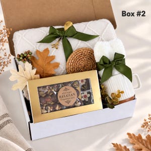 May include: A gift box labeled Box #2 containing a white textured blanket tied with a green ribbon, a round woven coaster, a white mug with a green ribbon, and a gold box of Artisan Belgian Chocolate with floral-topped chocolates.