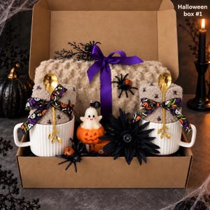 May include: A cardboard gift box labeled Halloween box #1 containing a beige textured blanket with a purple ribbon, two white ribbed mugs with gold spoons and patterned bows, a ghost on a pumpkin figurine, and black spider decorations.