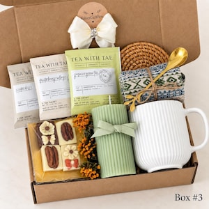 May include: A gift box containing three Tea With Tae tea sachets, a white ribbed ceramic mug, a sage green pillar candle with a ribbon, a gold spoon, a patterned fabric item, and a clear package of four square white chocolates with pecan toppings.