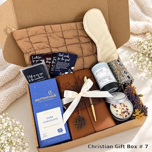 May include: A gift box containing a brown quilted pouch, a cream sleep mask, a blue Moonstruck dark chocolate bar, a brown journal with a leaf charm and white ribbon, a tea tin, a candle, a patterned cloth, and cards with religious text.