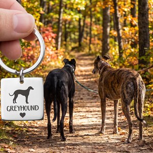 May include: A square silver-toned metal keychain featuring a black silhouette of a greyhound dog, the text "GREYHOUND" in a sans-serif font, and a small black heart symbol centered below the text, attached to a circular metal split ring.