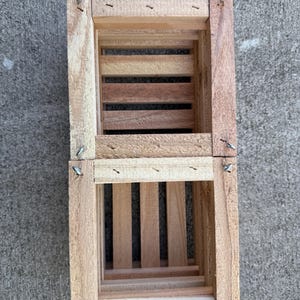 May include: Two small, rectangular wooden crates stacked on top of each other. The crates are constructed from light-colored wood with darker wood slats forming the base and sides. Metal staples secure the corners.