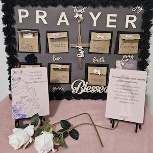 Puede incluir: Un tablero de oración rectangular con un borde floral negro, que presenta la palabra PRAYER en letras blancas, pequeños sobres marrones etiquetados por categorías, una cruz de madera y dos tarjetas con texto inspirador.