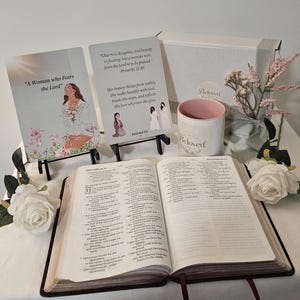 May include: An open Bible with two illustrated cards on black stands, a white mug with a pink interior labeled Beloved, and two white artificial roses. The cards feature text including A Woman who Fears the Lord and Proverbs 31:30.