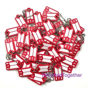 Könnte beinhalten: Eine Ansammlung von rechteckigen, rot-weißen Schlüsselanhängern mit dem BTS-Logo und Schriftzug. Jeder Anhänger ist an einer silberfarbenen Metallkugelkette befestigt. Die Artikel liegen in einem Haufen, und der Text NKraftedTogether ist lila.