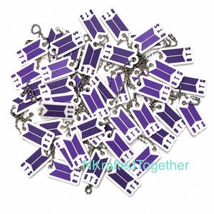 May include: A collection of rectangular white keychains featuring a purple BTS logo and the text BTS printed on each piece. Each keychain is attached to a silver-colored metal ball chain. The items are scattered in a pile on a white surface.