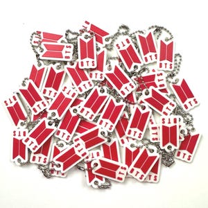 Pode incluir: Uma coleção de numerosos porta-chaves retangulares com um logótipo BTS vermelho e o texto BTS em branco. Cada porta-chaves está preso a uma corrente de bolas de metal prateado, criando um monte denso de artigos idênticos e uniformes.