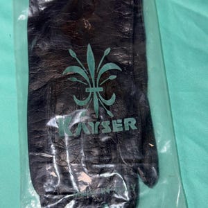 May include: A pair of black leather gloves enclosed in a clear plastic bag. The gloves feature a textured surface and a green logo printed on the front, displaying a fleur-de-lis symbol above the brand name KAYSER and the text MADE IN ITALY.