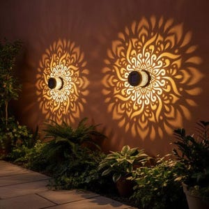 Puede incluir: Dos luces de pared solares proyectan intrincados patrones tipo mandala sobre una pared marrón. Las luces son circulares con un diseño semicircular, y los patrones se iluminan en un cálido tono dorado. Plantas verdes y follaje son visibles en primer plano.