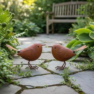 Könnte beinhalten: Zwei rostige, braune Metallvogel-Skulpturen stehen sich auf einem Steinweg gegenüber. Die Vögel haben lange Schwanzfedern und dünne Beine. Im Hintergrund sind Grün und eine Holzbank zu sehen.