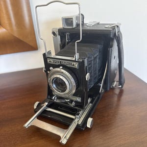 May include: A vintage Speed Graphic press camera featuring a black leather-covered body, a folding bellows mechanism, a silver metal wire frame viewfinder, and a circular lens assembly with visible aperture and shutter speed settings.