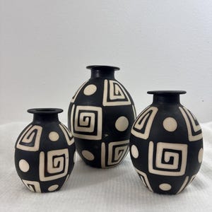 May include: A set of three matte black vases of varying sizes, each featuring a contrasting white geometric pattern consisting of repeating square spirals and solid circles. The vases have a rounded body shape with a narrow neck and a flat rim.