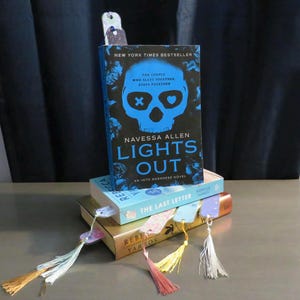 May include: A stack of books with colorful bookmarks. The top book is titled "LIGHTS OUT" by Navessa Allen, with a blue skull design. Other books include "THE LAST LETTER" and "REBECCA YARROS". The bookmarks have tassels.