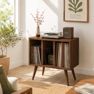 Puede incluir: Un mueble para tocadiscos de estilo moderno de mediados de siglo en madera marrón cálido. El mueble cuenta con un tocadiscos, un pequeño altavoz y espacio para guardar discos de vinilo. Una jarrón con flores rosas encima.