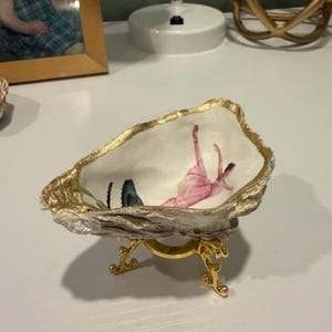 May include: A decorative oyster shell dish with a gold-painted rim, featuring an interior illustration of a pink-clad dancer and a blue butterfly. The shell is supported by a small, ornate gold-coloured metal tripod stand with curved legs.