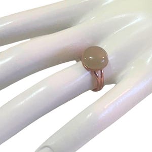 May include: A ring featuring a smooth, round, beige-colored cabochon stone set in a polished rose gold-toned metal band. The band has a split design that connects to the base of the circular stone setting, creating a simple and clean appearance.
