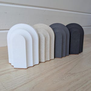 Puede incluir: Un conjunto de cuatro objetos decorativos en forma de arco en tonos blanco, beige, gris y negro. Cada pieza tiene un diseño en capas y se asienta sobre una superficie de madera. Los objetos están dispuestos en fila, mostrando una estética minimalista.