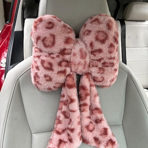 May include: A pink, plush car headrest cover shaped like a bow. The cover features a leopard print pattern in a darker shade of pink. The headrest is shown installed on a light gray car seat.
