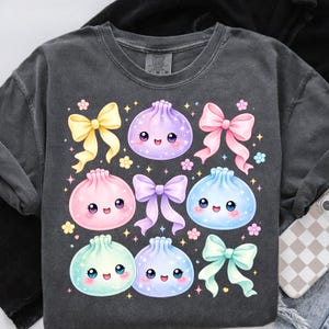 May include: A charcoal gray t-shirt featuring a graphic print of five pastel-colored, smiling dumpling characters interspersed with yellow, pink, purple, and mint green bows, accented by small sparkling stars and tiny flower shapes.