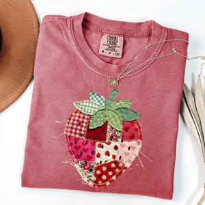 Puede incluir: Una camiseta rosa polvoriento con una gran aplicación central de fresa hecha de varios cuadrados de tela de patchwork en tonos rojos, rosas y blancos con patrones florales y de corazones. La camiseta tiene una etiqueta de la marca Comfort Colors.
