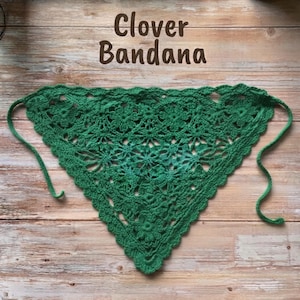 Könnte beinhalten: Ein dreieckiges grünes Häkelbandana mit einem sich wiederholenden Blumenmuster, gewellten Rändern und dünnen Bändern an jeder Seite. Der Text Clover Bandana ist in dunkelbraunen Buchstaben über dem grünen Häkelstoffstück aufgedruckt.