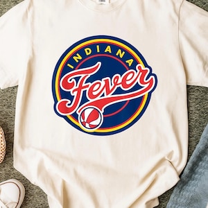 May include: A cream-colored short-sleeve t-shirt featuring a circular graphic with a dark blue background, yellow and red borders, and the text "INDIANA Fever" in red script. A small basketball graphic is positioned below the word Fever.