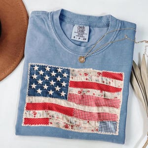 Op de afbeelding: Een blauw t-shirt met ronde hals, een Comfort Colors label en maat S. De voorzijde is voorzien van een rechthoekige patch in de vorm van de Amerikaanse vlag, samengesteld uit diverse stoffen zoals rode stippen, bloemenprints en vichy-ruitjes.