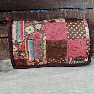 May include: A rectangular quilted fabric pouch featuring a patchwork design of pink, brown, and cream floral, polka dot, and striped patterns. The pouch has a dark brown fabric border, a zipper closure, and a small oval metal tag on the bottom right.