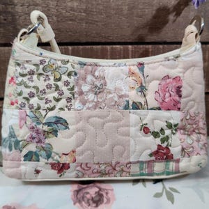 May include: A small, quilted handbag with a patchwork design. The bag features a variety of floral patterns in shades of pink, cream, and green. It has a curved shape, a cream-colored strap, and silver metal rings.
