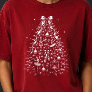 May include: A dark red t-shirt featuring a white graphic of a Christmas tree composed of ornaments, a nutcracker, ballet dancers, and ballet slippers. A large white ribbon bow sits at the top of the tree, with sparkling accents throughout the design.